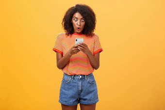 室内拍摄震惊了惊呆了非洲美国年轻的女人非洲式发型发型盯着惊讶兴奋智能手机屏幕持有手机阅读令人惊异的消息橙色墙