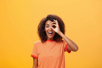 有创意的好玩的梦幻皮肤黝黑的成人女孩卷曲的发型条纹t恤显示圆眼睛手势微笑广泛的准备好了部分冒险橙色墙