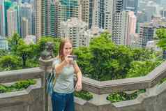 在香港香港维多利亚峰女人采取自拍坚持图片照片智能手机享受视图维多利亚港口查看平台前峰塔散焦背景旅行亚洲概念