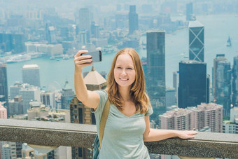 在香港香港维多利亚峰女人采取自拍坚持图片照片智能手机享受视图维多利亚港口查看平台前峰塔散焦背景旅行亚洲概念