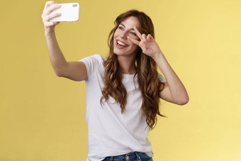快乐的有魅力的好看的卷发女人白色t恤显示和平胜利标志微笑广泛的摆姿势温柔的友好的表达式智能手机前面相机自拍黄色的背景