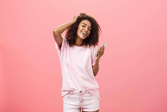 肖像无忧无虑的时尚的现代皮肤黝黑的年轻的女孩智能手机挂快乐触碰头发盯着广泛的微笑相机持有手机摆姿势粉红色的墙