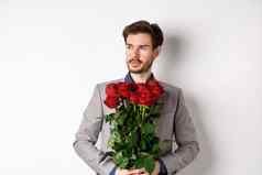 英俊的的男朋友西装浪漫的日期持有花束红色的玫瑰左深思熟虑的站白色背景