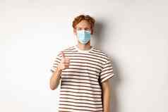 科维德流感大流行社会距离概念年轻的男人。红色的头发穿医疗面具防止捕捉冠状病毒显示竖起大拇指白色背景