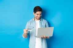 年轻的微笑男人。购买互联网持有信贷卡支付购买移动PC站蓝色的背景