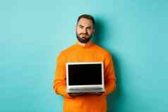 英俊的有胡子的男人。橙色毛衣显示移动PC屏幕展示促销扮鬼脸失望心烦意乱站光蓝色的背景