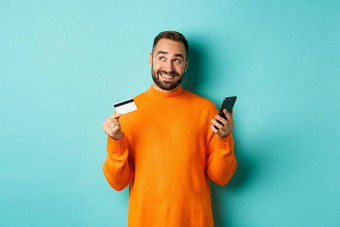 在线购物英俊的男人。思考持有智能手机信贷卡支付互联网商店站光蓝色的背景