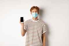 科维德流感大流行社会距离概念英俊的年轻的男人。脸面具显示黑色的智能手机屏幕微笑站白色背景