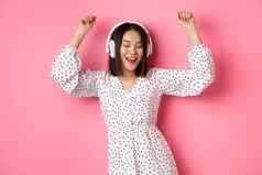 快乐年轻的亚洲女人跳舞有趣的听音乐耳机站粉红色的背景