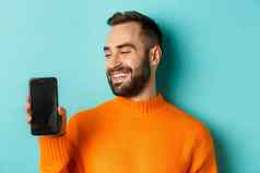 特写镜头年轻的有胡子的男人。显示电话屏幕满意穿橙色毛衣站工作室背景