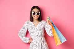 有吸引力的微笑亚洲女人持有购物袋穿太阳镜可爱的衣服站粉红色的背景