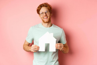 真正的房地产快乐的年轻的男人。红色的头发穿眼镜t恤显示纸房子断路微笑购买公寓粉红色的背景