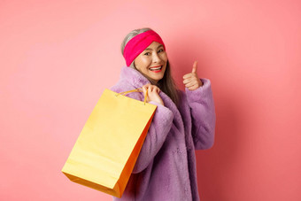 时尚的亚洲高级女人购物携带纸袋肩膀显示翘拇指推荐商店折扣粉红色的背景