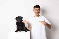 快乐的年轻的男人。指出手指狗显示小可爱的黑色的哈巴狗坐着白色背景