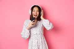 可爱的亚洲女人玩卡拉ok应用程序唱歌移动电话耳机站衣服粉红色的背景