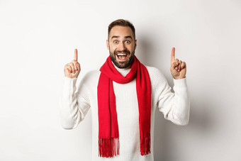 圣诞节购物冬天假期概念快乐的英俊的男人。显示促销横幅指出手指复制空间穿红色的围巾毛衣白色背景