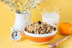 健康的丰盛的早餐烤格兰诺拉麦片燕麦坚果葡萄干碗玻璃牛奶橙色报警时钟黄色的背景