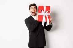 图像快乐好看的男人。黑色的西装持有盒子圣诞节礼物微笑站白色背景