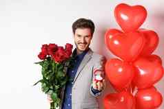 英俊的的男朋友西装使婚礼建议显示订婚环结婚持有红色的玫瑰站情人节一天气球白色背景