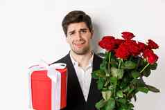 特写镜头持怀疑态度男人。西装持有花束红色的玫瑰礼物站不情愿的白色背景