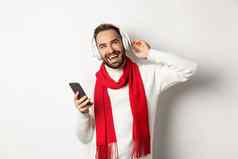 冬天假期技术概念男人。享受听音乐耳机满意持有智能手机穿毛衣围巾白色背景
