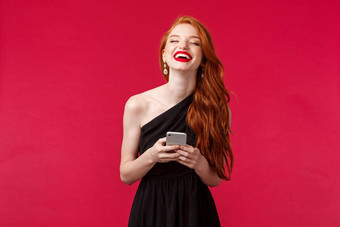 技术生活方式概念华丽的年轻的红色头发的人女人黑色的衣服笑红色的背景持有智能手机发短信朋友毕业舞会讨论有趣的时刻