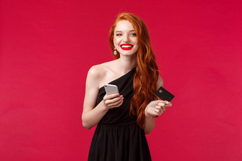 肖像迷人的年轻的红色头发的人女孩黑色的衣服长姜头发持有信贷卡智能<strong>手机</strong>支付在线购买<strong>订单</strong>装聚会，派对站红色的背景