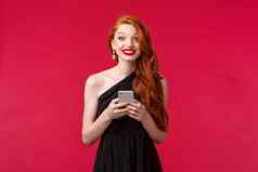 肖像兴奋逗乐红色头发的人女人穿黑色的衣服日期毕业舞会晚上持有移动电话微笑喜气洋洋的相机站红色的背景乐观的
