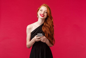 肖像华丽的微笑女人姜头发优雅的黑色的毕业舞会衣服持有智能手机消息传递应用程序图片笑相机红色的背景
