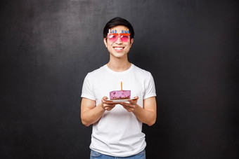 生日庆祝活动聚会，派对概念快乐年轻的快乐的亚洲的家伙白色t恤有趣的眼镜持有生日蛋糕蜡烛打击使微笑兴奋站黑色的背景