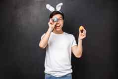 假期聚会，派对复活节概念逗乐可爱的亚洲愚蠢的的家伙兔子耳朵眼镜简单的t恤持有画鸡蛋眨眼隐藏眼睛开玩笑地站黑色的背景