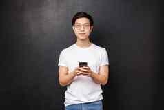 技术小工具人概念英俊的年轻的确定亚洲的家伙t恤眼镜移动电话应用程序持有智能手机微笑相机黑色的背景