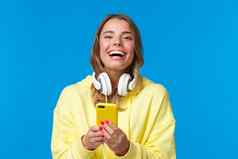 特写镜头肖像快乐的好看的金发碧眼的女孩黄色的连帽衫白色耳机移动电话写作博客分享新闻社会媒体消息传递笑相机