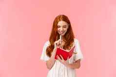 深思熟虑的快乐微笑漂亮的红色头发的人女白色衣服触碰下巴笔思考使杂货店待办事项列表写作可爱的红色的笔记本粉红色的背景