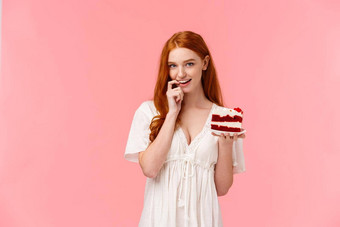诱人的卖弄风情的红色头发的人诱人的女朋友建议和平蛋糕持有美味的甜点咬手指微笑轻浮相机站粉红色的背景抵制诱惑