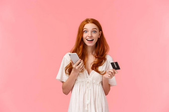 深刻的印象兴奋快乐红色头发的人女孩白色衣服惊讶<strong>容易</strong>使购买持有智能手机信贷卡微笑逗乐想知道相机粉红色的背景