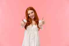 支持选择有魅力的兴奋快乐红色头发的人女人白色衣服显示竖起大拇指欢呼同意表达满意度很酷的产品微笑批准粉红色的背景