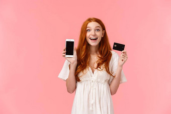 诱人的很高兴着迷可爱的快乐红色头发的人女孩微笑满意逗乐持有信贷卡显示智能手机显示使伟大的购买吹牛朋友粉红色的背景