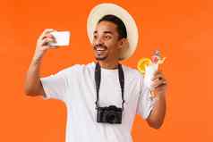 快乐的有魅力的英俊的非裔美国人男人。他相机白色t恤提高玻璃鸡尾酒享受喝让人耳目一新海边采取自拍假期橙色背景