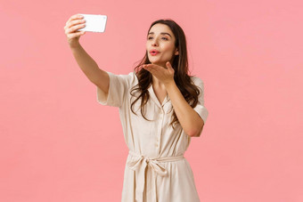 可爱的温柔的诱人的浅黑肤色的女人女衣服持有智能手机采取自拍吹吻移动相机流生活视频博客拍摄粉红色的背景
