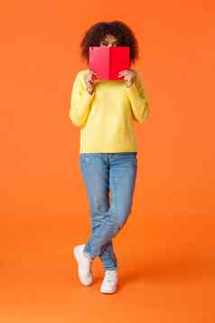 全身的垂直拍摄害羞的可爱的非裔美国人现代城市女孩非洲式发型发型隐藏脸红色的笔记本窥视相机写作日记站橙色背景