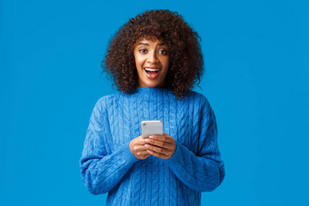 兴奋不知所措微笑有吸引力的年轻的非裔美国人女人穿冬天蓝色的毛衣惊讶惊讶相机收到伟大的新闻智能手机消息