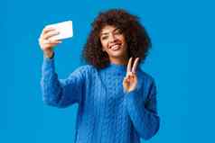 快乐的友好的可爱的非裔美国人女学生采取照片应用过滤器智能手机应用程序采取自拍倾斜头可爱的微笑使和平手势蓝色的背景