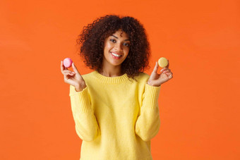 半身照肖像可爱的年轻的非裔美国人女卷曲的非洲式发型发型持有马卡龙微笑吃糖果讨厌饮食糕点甜点站橙色背景