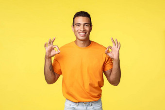 迷人的快乐的家伙纹身手臂橙色t恤白色裤子显示确认好手势微笑点头协议推荐产品促进公司黄色的背景