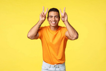 快乐可爱的可爱的拉美裔的男朋友橙色t恤显示和平胜利手势头微笑愚蠢的傻瓜使温柔的表达式使女朋友笑黄色的背景