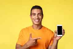 英俊的愉快的年轻的男人。橙色t恤持有智能手机指出移动显示微笑满意推荐应用程序显示促销活动电话应用程序吹牛买了在线