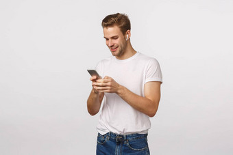微笑快乐金发碧眼的欧洲的家伙白色t恤牛仔裤持有智能手机穿无线耳机移动显示阅读消息选择首歌白色背景