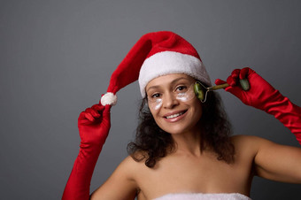 年轻的美丽的女人穿着圣诞老人玉辊按摩器淋巴排水面部按摩提出了灰色的背景复制空间圣诞节广告美客厅皮肤护理