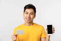 英俊的可爱的微笑亚洲男人。黄色的t恤显示智能手机显示信贷卡咧着嘴笑很高兴推荐银行服务在线支付系统站白色背景